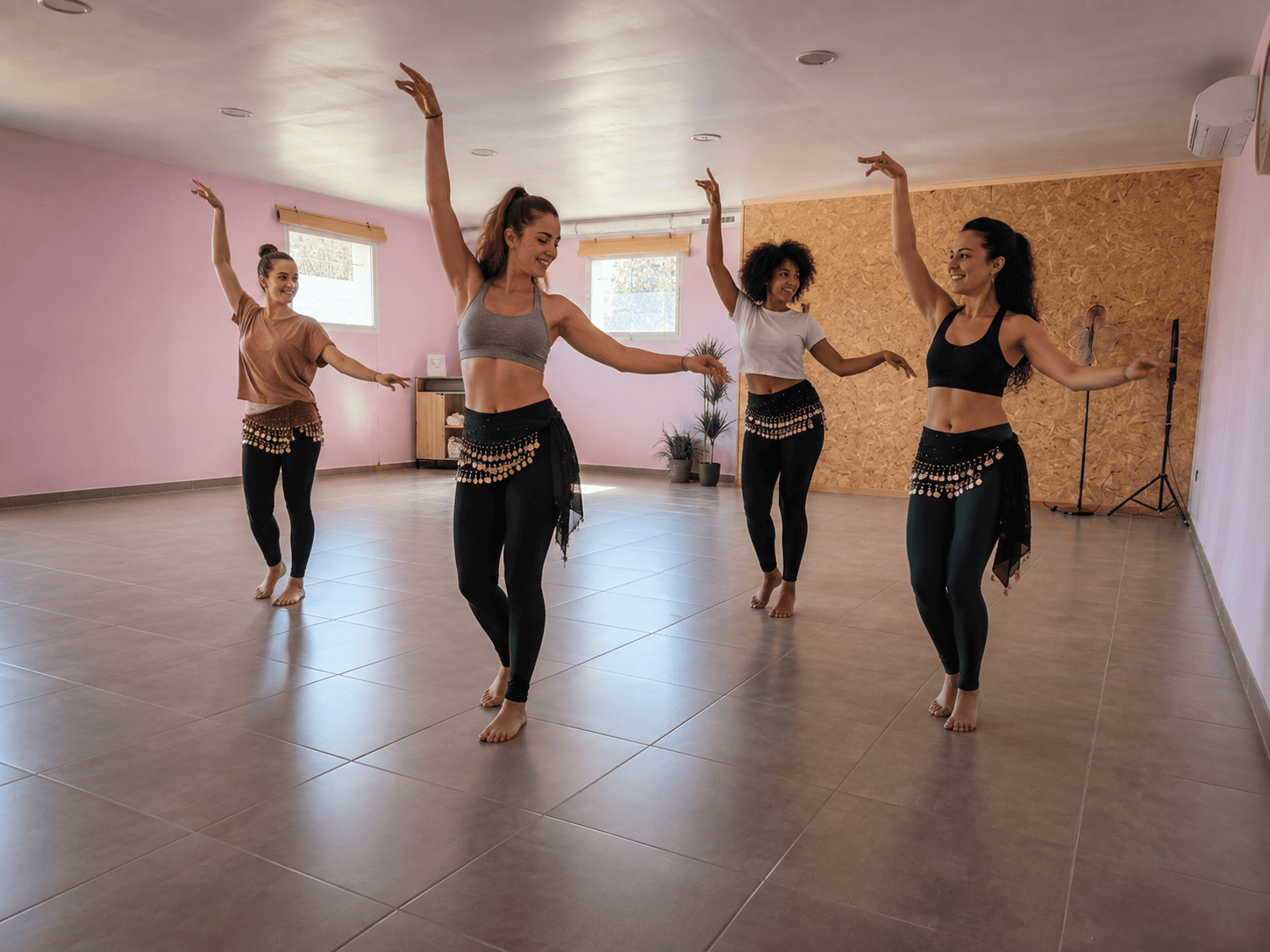 Quatre femmes en tenue de sport pratiquent la danse orientale dans une salle de fitness lumineuse, avec des foulards de danse autour des hanches.